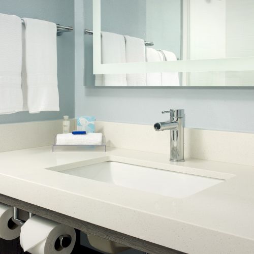 A bathroom vanity with a sink, faucet, mirror, two white towels on a rack, toiletries, and toilet paper under the counter.