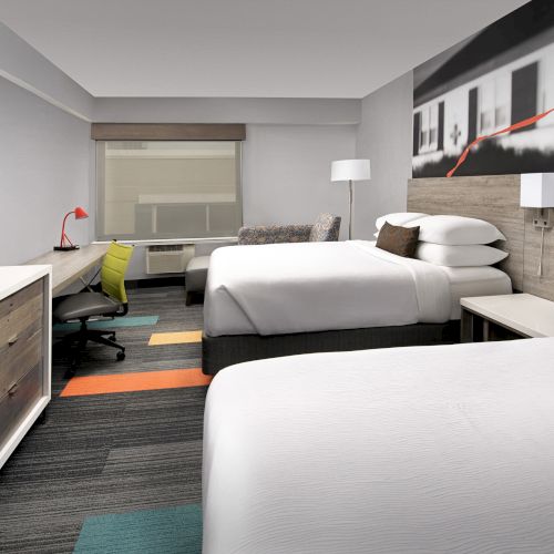 The image shows a modern hotel room with two beds, wall art, a TV, desk, chair, and lamp, featuring a bright and contemporary design.
