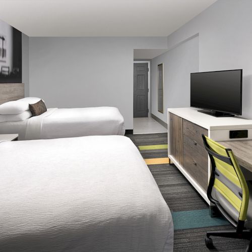 The image shows a modern hotel room with two beds, a desk with a red lamp, a flat-screen TV, and minimalist decor.
