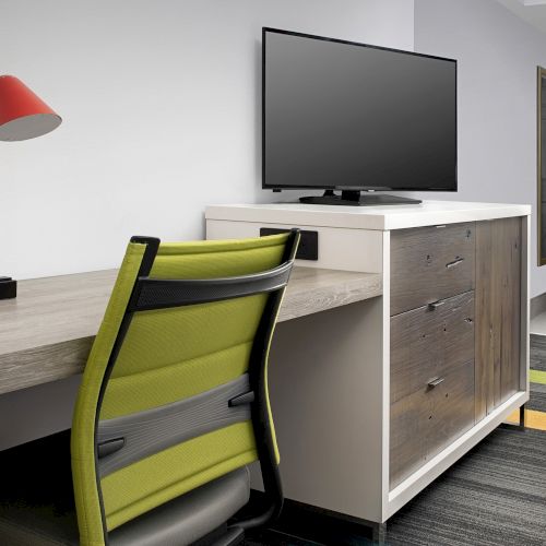 The image shows a modern office setup with a desk, red lamp, green chair, and a TV on a cabinet.