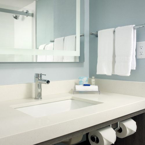 A modern bathroom with a rectangular sink, a mirror, towels on a rack, and toiletries on the counter. Toilet paper is beneath the counter.
