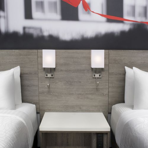 The image shows a modern hotel room with two beds, a central nightstand, and wall lights, featuring a red decorative element above the beds.