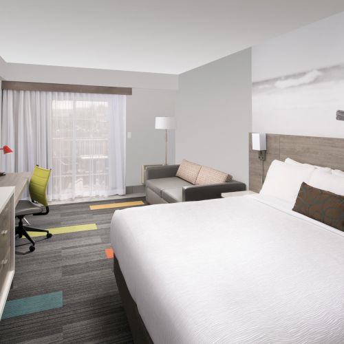 A modern hotel room with a large bed, sofa, desk, TV, and wide window with curtains, featuring neutral and orange accents.