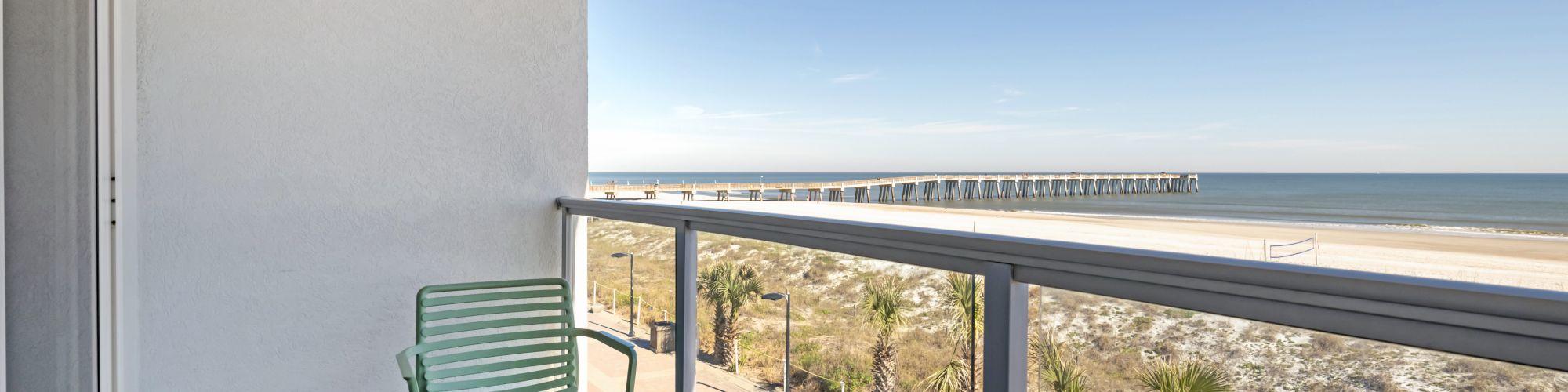 A balcony with two green chairs and a small table overlooks a beach and pier under a clear blue sky and open ocean view.
