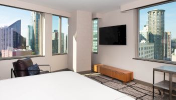 A modern hotel room with large windows offers city views, featuring a bed, wall-mounted TV, and contemporary furniture.
