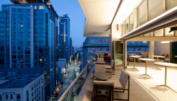 A modern rooftop terrace with seating, overlooking a cityscape of tall buildings and a street lit by evening lights.