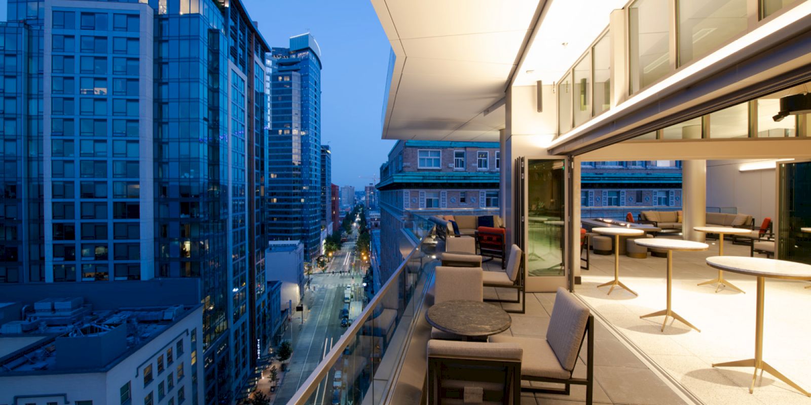 A rooftop patio with seating overlooks a city street lined with modern buildings, captured during twilight.