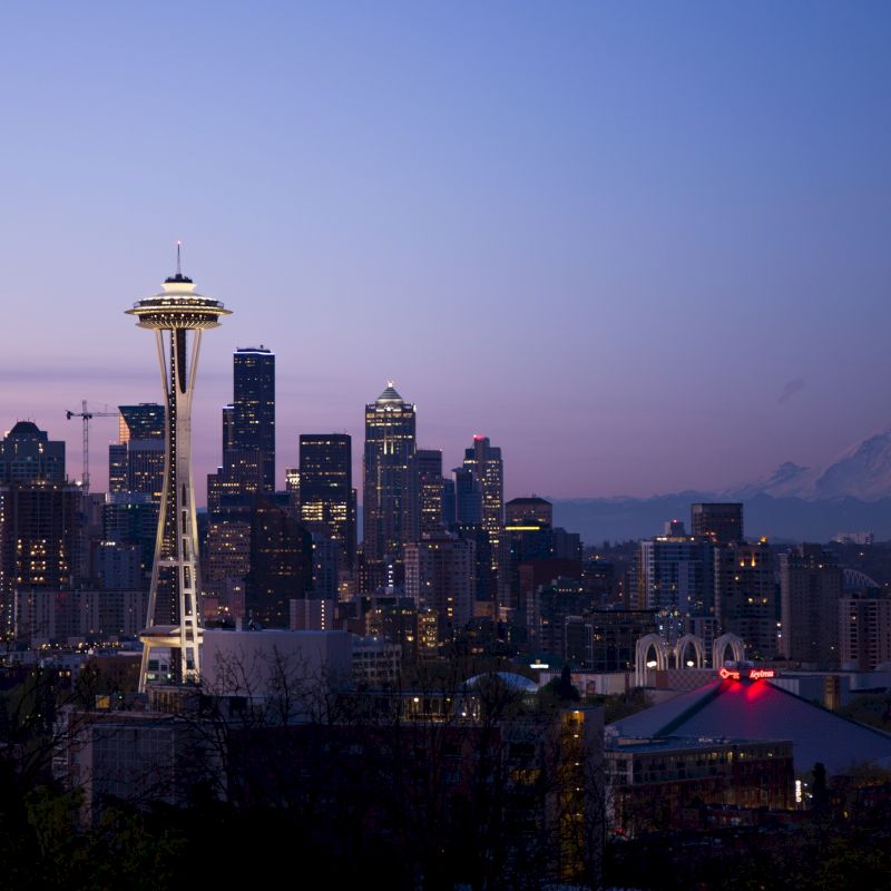 Seattle skyline at dusk featuring the Space Needle, with Mount Rainier in the background against a purple-blue sky.