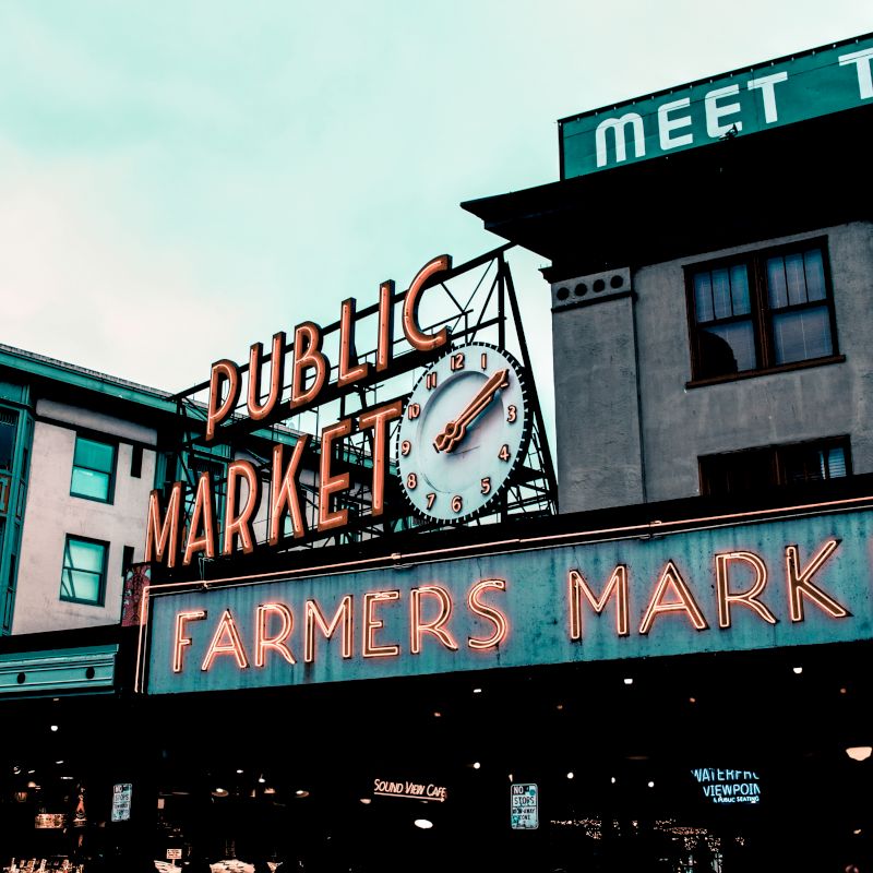The image shows the entrance to a public and farmers market with a vintage neon sign and clock, set against an urban building backdrop.