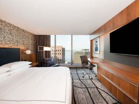 A modern hotel room with a large bed, patterned wall, TV, desk, and window offering a city view, providing a comfortable stay.