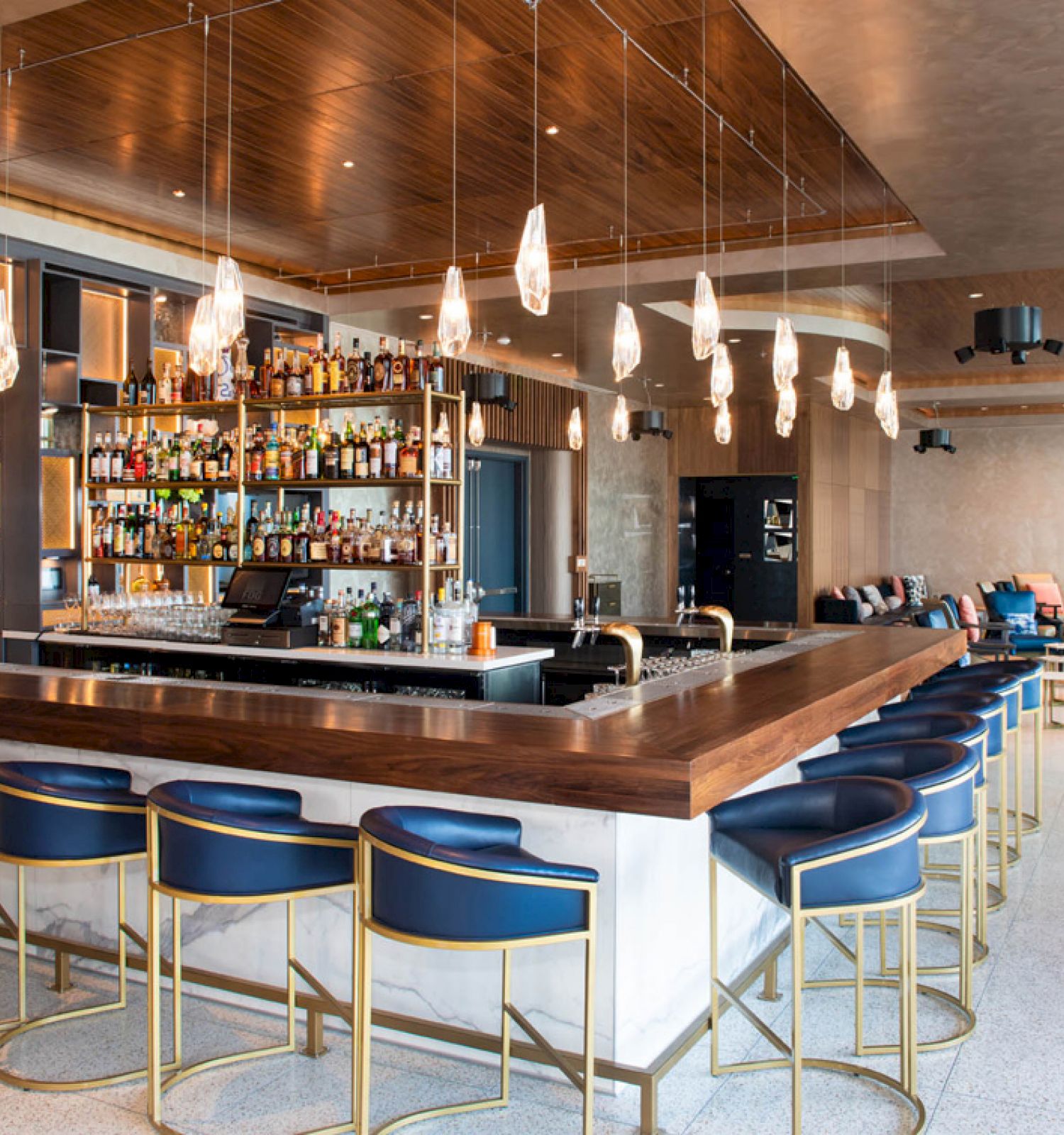 The image shows a modern bar with a wooden counter, blue stools, pendant lights, and shelves stocked with bottles.