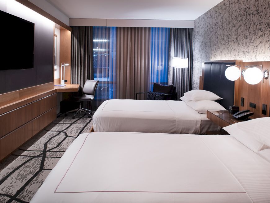 The image shows a hotel room with two beds, modern decor, a wall-mounted TV, desk, lamps, and a window with curtains.