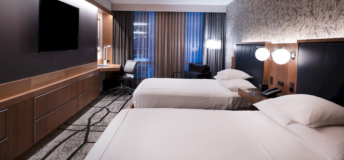 The image shows a hotel room with two beds, modern decor, a wall-mounted TV, desk, lamps, and a window with curtains.