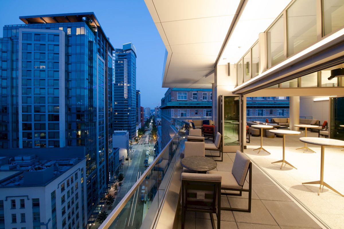 The image shows a modern rooftop terrace with seating, overlooking a city street lined with tall buildings, during the evening.