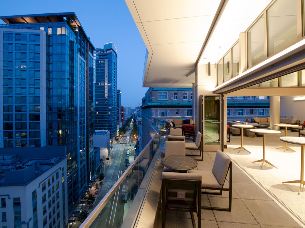 The image shows a modern rooftop terrace with seating, overlooking a city street lined with tall buildings, during the evening.
