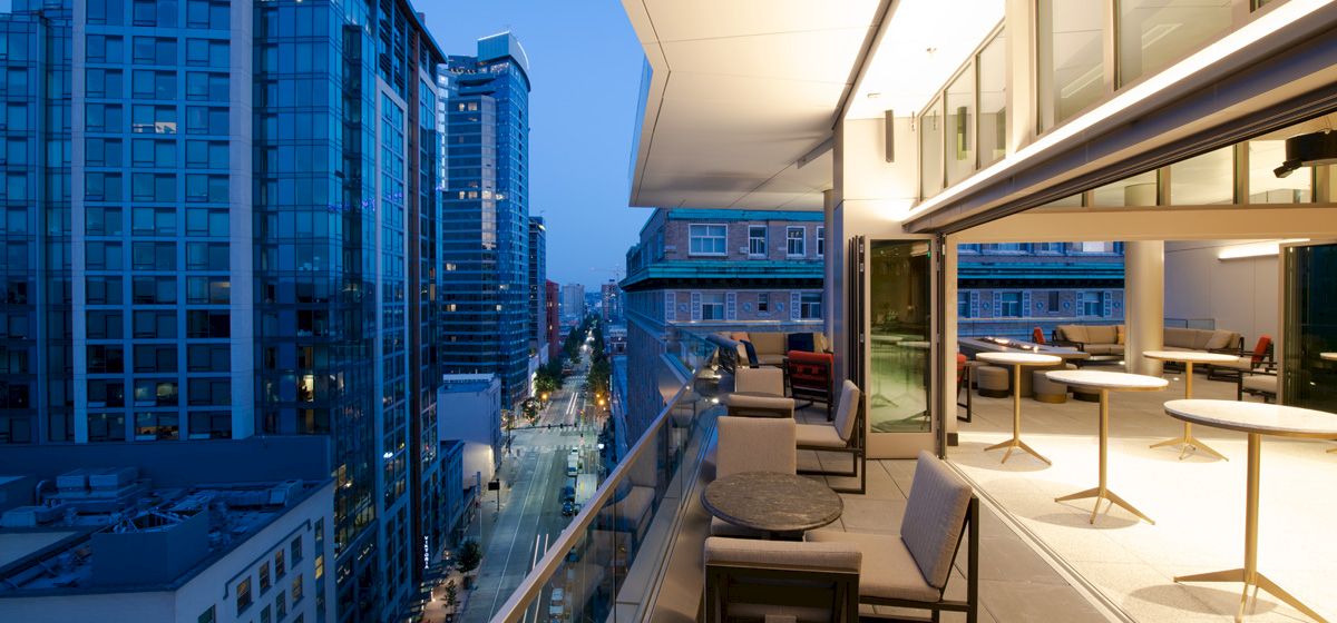 The image shows a modern rooftop terrace with seating, overlooking a city street lined with tall buildings, during the evening.
