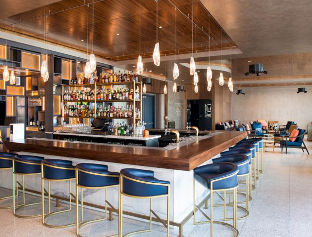 A modern bar with a sleek countertop, blue bar stools, pendant lighting, and shelves stocked with bottles; seating area in the background.