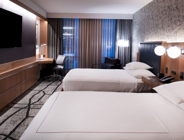 The image shows a modern hotel room with two beds, a flat-screen TV, a desk, and large windows with curtains, creating a cozy and stylish ambiance.