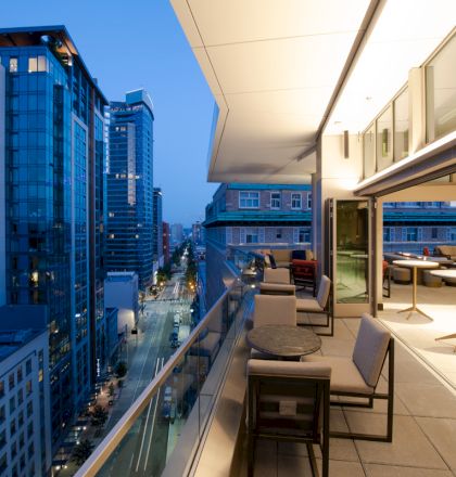 A rooftop terrace with seating overlooks a cityscape of tall buildings at dusk, featuring a modern and vibrant urban environment.