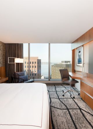 A modern hotel room with a large bed, wall-mounted TV, desk, chair, and window with a city view.