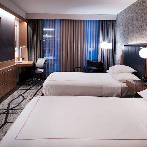 A modern hotel room with two beds, a desk, a large TV, and ambient lighting.