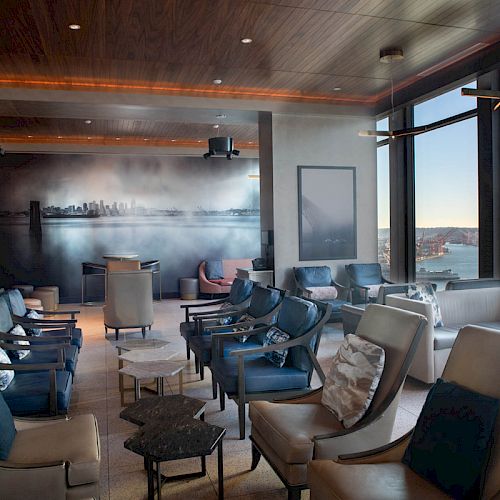 A modern lounge with comfortable seating, large windows displaying a scenic view, and a wall mural depicting a city skyline.