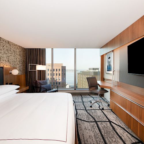 A modern hotel room features a large bed, wall-mounted TV, desk area, and a window with a city view.