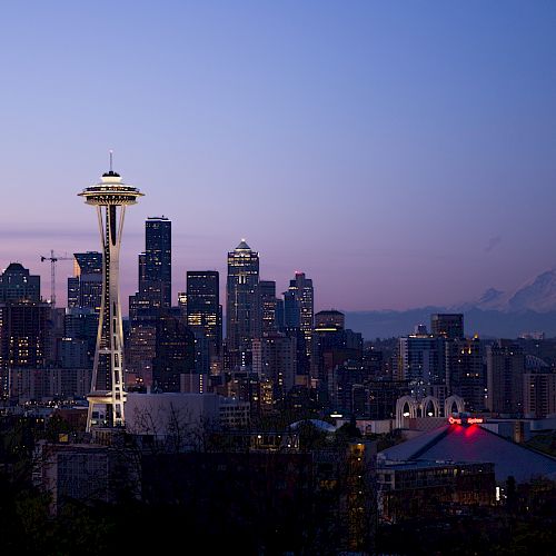 City skyline at dusk with a prominent observation tower and mountains in the background, glowing lights.