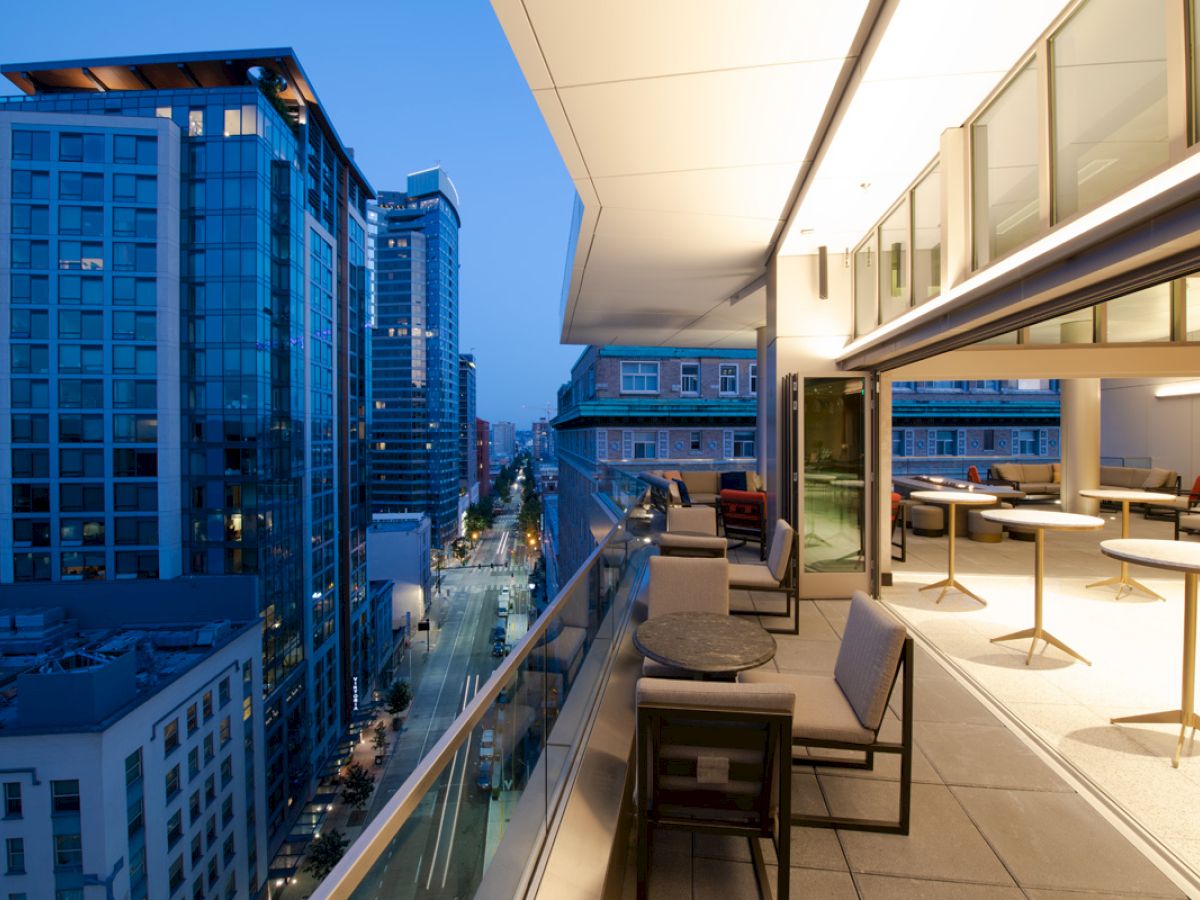 A modern rooftop terrace with seating overlooks a cityscape featuring tall buildings and a street lit up at dusk.