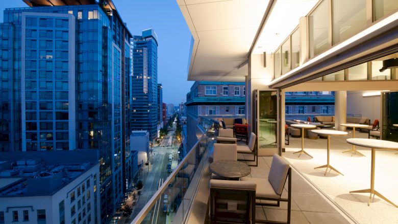A modern rooftop terrace with seating overlooks a cityscape featuring tall buildings and a street lit up at dusk.