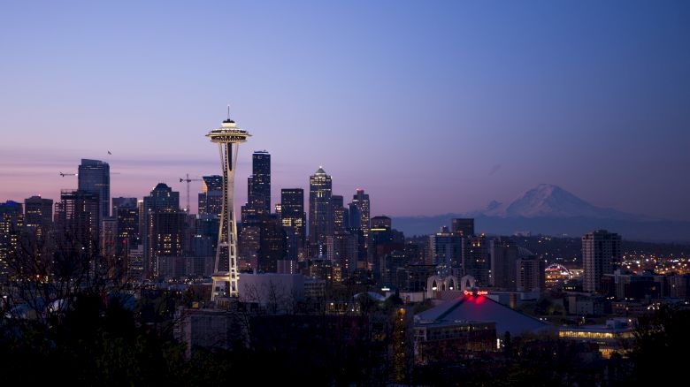 The image shows the Seattle skyline at dusk, with the Space Needle prominently visible and a mountain in the background.