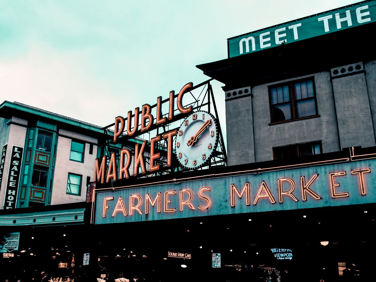 The image shows a neon sign of a public farmers market with a clock, set against urban buildings under a cloudy sky.