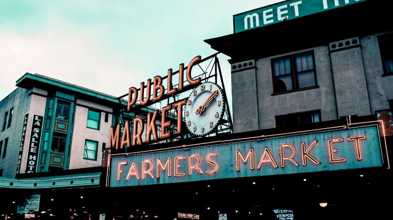 The image shows a neon sign of a public farmers market with a clock, set against urban buildings under a cloudy sky.