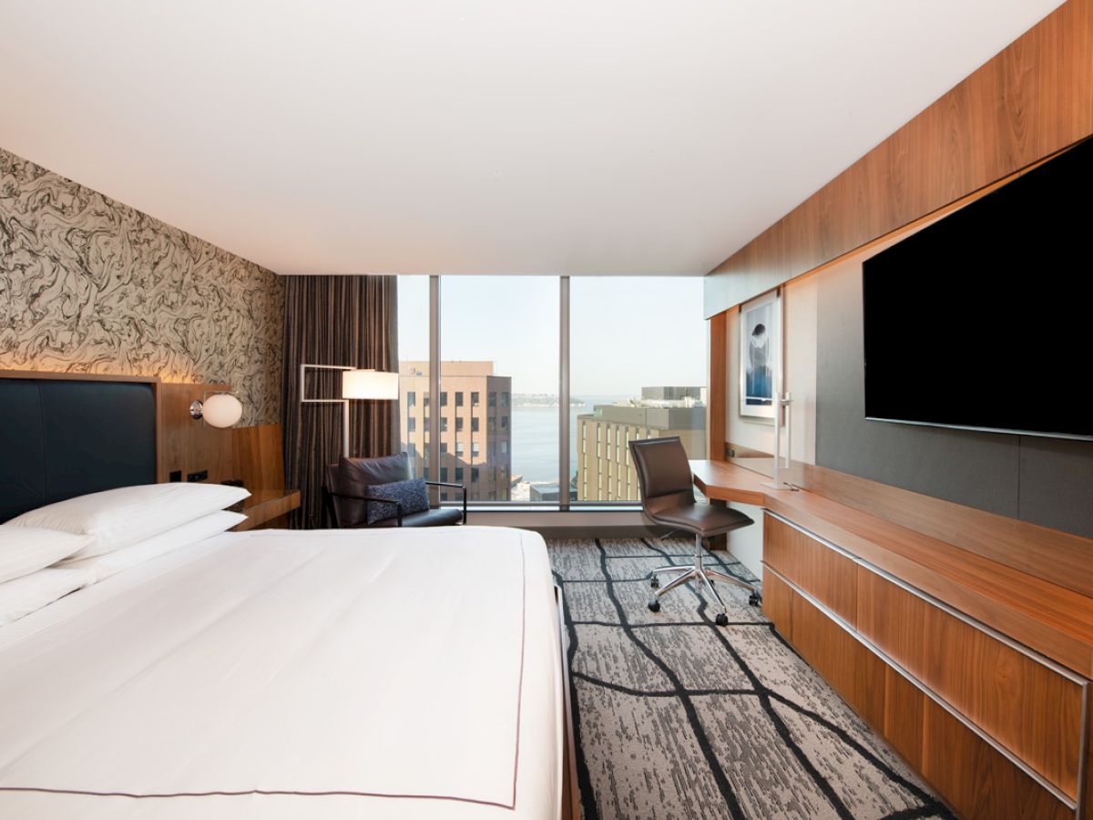 A modern hotel room with a large bed, desk, chair, and a widescreen TV, featuring a city view through a large window.