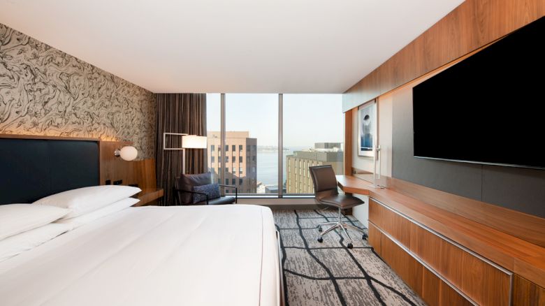 A modern hotel room with a large bed, desk, chair, and a widescreen TV, featuring a city view through a large window.