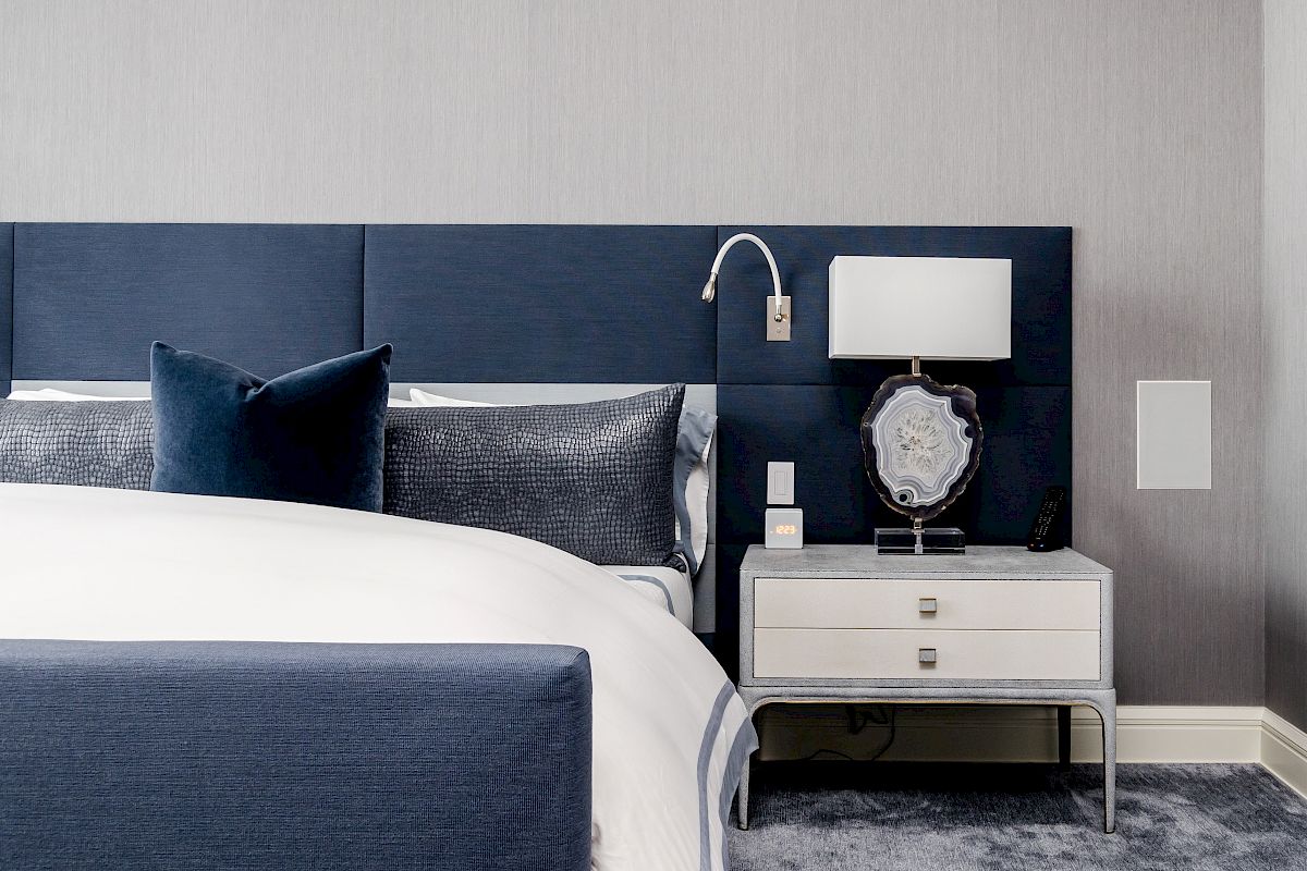 A modern bedroom with a navy headboard, white nightstand, and stylish lamp on a light carpet. It looks cozy and contemporary.