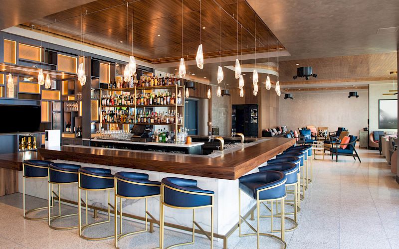 The image shows a modern bar with blue stools, hanging lights, and a fully stocked counter in a stylish, spacious setting.