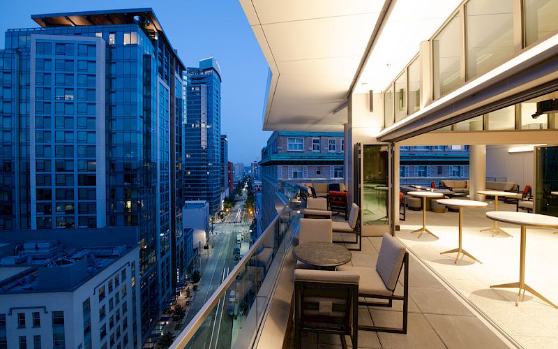 A modern rooftop terrace at dusk, overlooking a cityscape with tall buildings, outdoor seating, and warm lighting creating a cozy ambiance.