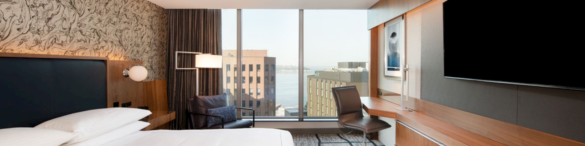 A modern hotel room with a bed, large TV, desk, and chair; a cityscape view is seen through the floor-to-ceiling window.